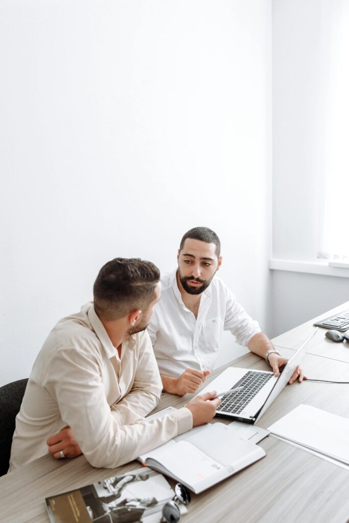 Two businessmen in white shirts engaged in a discussion over a laptop in a bright office setting.